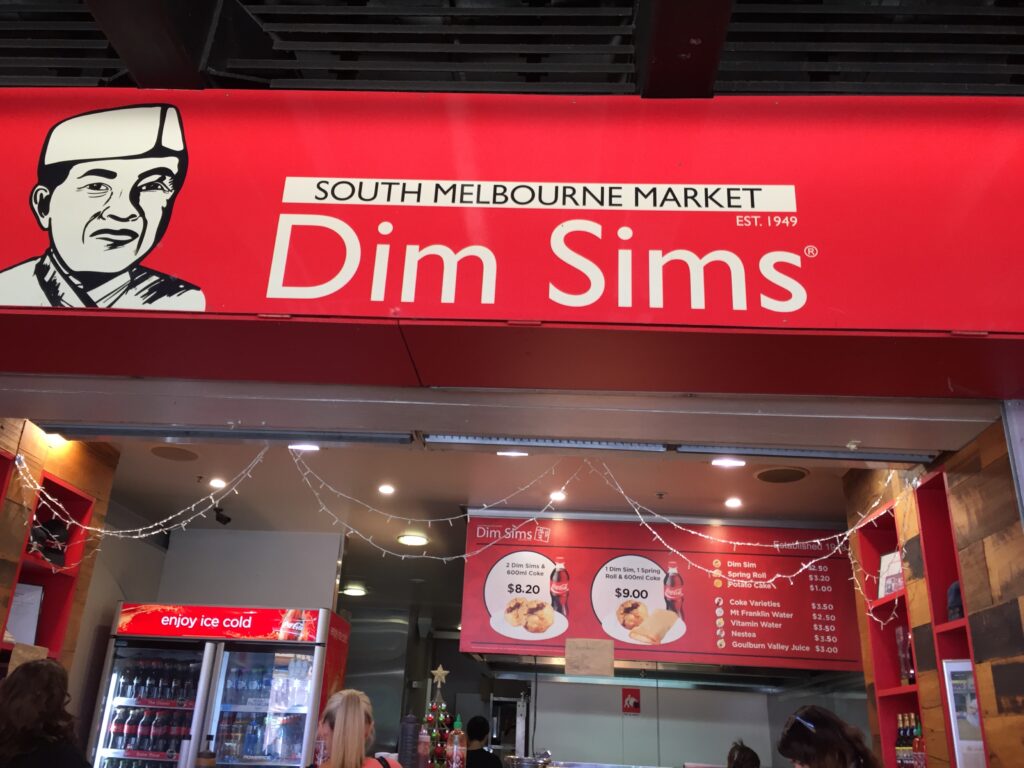 The front of the South Melbourne Market Dim Sims shop, featuring a bright red sign with a black-and-white illustration of a man in a chef’s hat and the words “South Melbourne Market Dim Sims Est. 1949.” Below, the counter displays combo meal prices alongside a fridge with drinks and a menu board. Customers are visible ordering at the counter.
